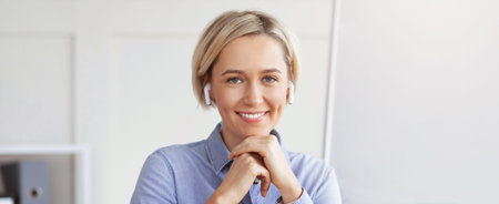 Woman Smiles While Sitting at a Desk With Earbuds in a Bright Office Space During the Dayの写真素材
