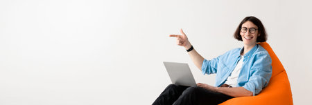 Person Sitting on a Bean Bag Chair Using a Laptop and Pointing With a Smile in a Simple Roomの写真素材