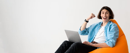 Young Man Sits on Bean Bag Chair and Works on Laptop With Happy Expression in Bright Roomの写真素材