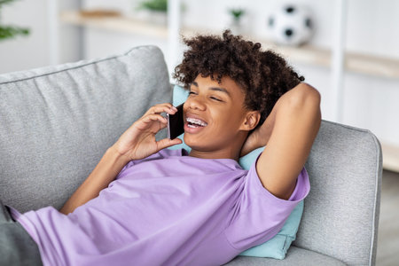 Cheerful black teenager lying on couch, having smartphone conversation with friend at home