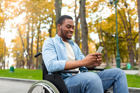 Cheery black guy in wheelchair using mobile phone, checking email or messages at autumn parkの写真素材