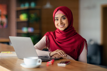 Cheerful muslim woman using modern laptop, cafe interiorの写真素材