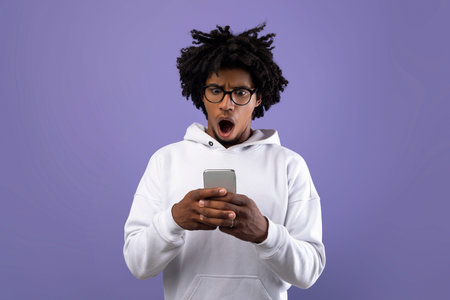 Frustrated black teen guy staring at smartphone in shock over violet studio background