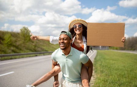 Happy young multiracial couple hitchhiking together, holding empty cardboard sign, showing thumb up gesture outdoorsの写真素材