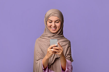 Excited young muslim woman in hijab using smartphone, texting or browsing internet, purple...