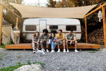 Group of cheery young diverse people with their cute dog sitting near motorhome, having fun camping...