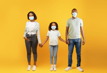 Young black parents and their child daughter wearing medical face masks and holding hands, yellow...