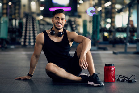 Arab Male Athlete Sitting At Gym Next To Container With Whey Protein