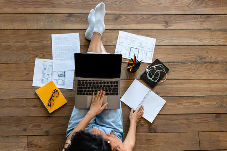Top view of woman with work or study materials using laptop with empty screen while sitting on floor...