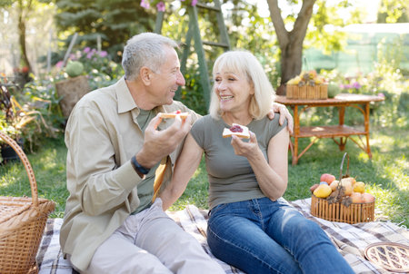Happy aged couple having picnic in their garden, sitting on blanket and eating toasts with jam, enjoying spring dayの写真素材