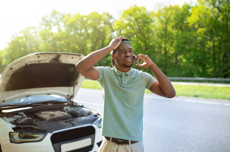 Black man standing near his broken car with open hood, calling breakdown road service on smartphone