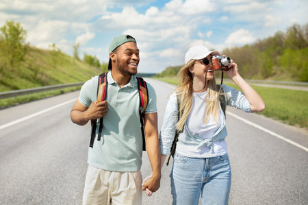 Happy young multiracial couple hitchhiking together, holding hands, taking photos on camera, walking...