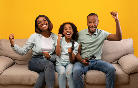Family time activities. Happy black parents and daughter playing video games, sitting on sofa over yellow backgroundの写真素材