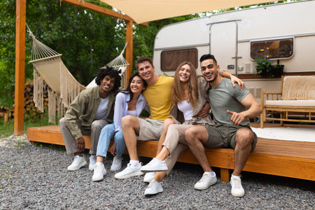 Full length portrait of multiracial young friends sitting near camper van, smiling at camera...