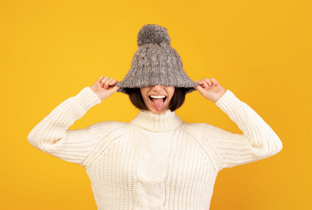 Playful millennial woman pulling down woollen hat and sticking out her tongue, fooling over yellow...