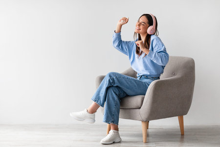 Happy millennial lady in wireless headphones relaxing in armchair and enjoying music against white studio wallの写真素材