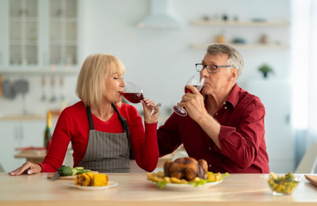 Affectionate senior couple drinking red wine while preparing festive Christmas dinner, making roasted turkey at kitchenの写真素材