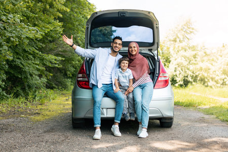 Positive auto journey. Happy millennial arabic guy and lady in hijab with baby, sit in car trunk and gesturingの写真素材