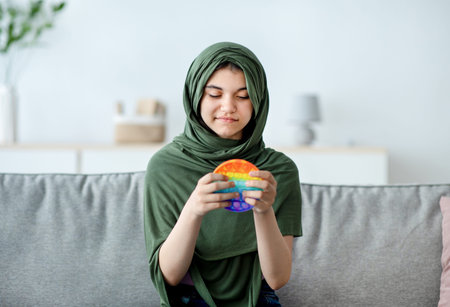 Portrait of lovely Indian teen girl in hijab playing with bright POP IT toy, enjoying fun antistress game at homeの写真素材