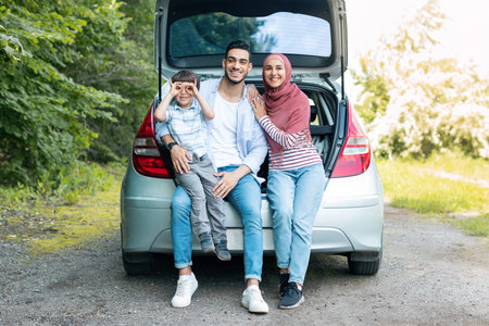Smiling young middle eastern man and wife in hijab holding baby, sitting in car trunk, resting from trip outdoorの写真素材