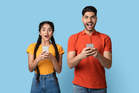 Excited Young Adults Using Smartphones Against a Light Blue Background While Enjoying a Fun Moment...