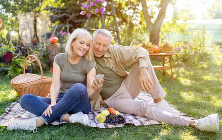 Happy elderly couple relaxing in garden and using smartphone, sitting on blanket on grass, resting outdoors, copy spaceの写真素材