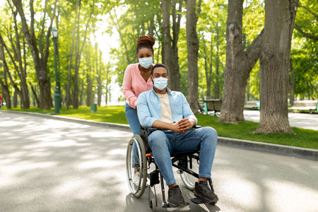 Full length of millennial black woman on walk with her impaired husband in wheelchair, wearing face...