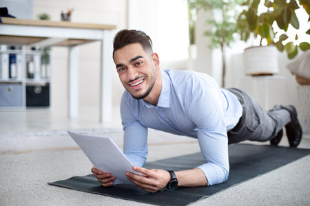 Workplace fitness. Happy Arab employee exercising on sports mat, standing in elbow plank with business papers at officeの写真素材