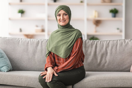 Happy Mature Muslim Woman Wearing Hijab Sitting On Sofa Indoors