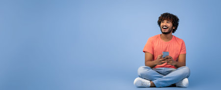 Man Sitting on Floor With Phone Smiling Against Plain Blue Backgroundの写真素材