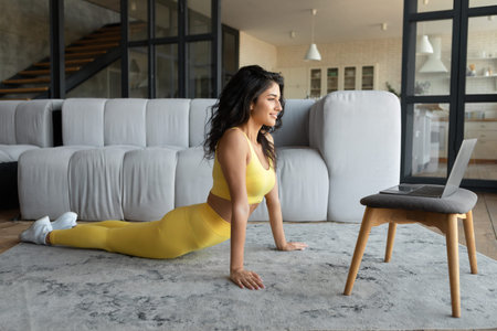 Happy young woman practicing yoga, making cobra pose in front of laptop at home, full length