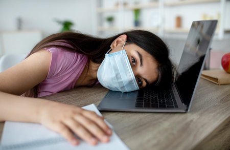 Unhappy Indian teen girl in face mask lying on laptop keyboard, feeling bored of online studies...