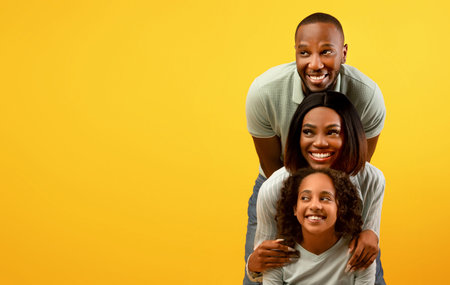 Positive african american family of three looking aside at empty space on yellow background, standing behind each otherの写真素材