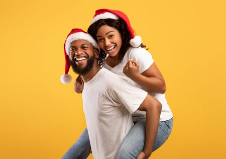 Happy black guy giving his girlfriend piggyback ride, wearing together Santa hats, having fun over yellow backgroundの写真素材