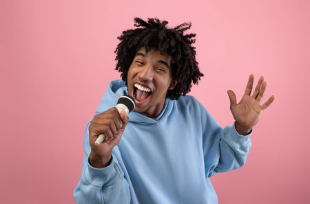 Portrait of black teen guy singing song, using microphone, performing karaoke on pink background