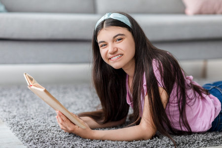 Domestic hobbies concept. Cheerful Indian teenage girl reading exciting book, lying on soft carpet at homeの写真素材