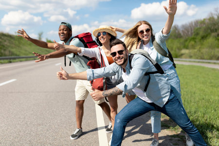 Cheerful young people standing on roadside, stopping car, trying to get free ride, having autostop journey, free spaceの写真素材