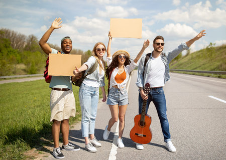 Happy diverse friends standing on highway with cardboard sign, waving down car, going on journey together, outdoorsの写真素材