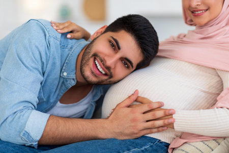 Future father. Excited muslim man listening his pregnant wifes belly, loving arab guy embracing big tummy, closeupの写真素材