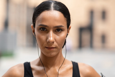 Confident millennial arab female athlete brunette in headphones look at camera on building backgroundの写真素材