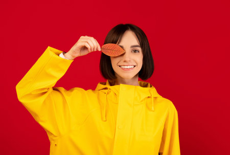 Happy young lady closing her eye with autumn leaf, posing and smiling to camera over red backgroundの写真素材