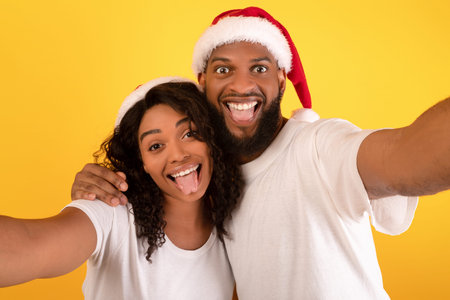 Playful black spouses in Santa hats taking selfie on yellow background