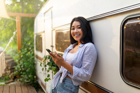 Happy Asian woman standing near camper van, using smartphone on camping trip in countryside, copy...