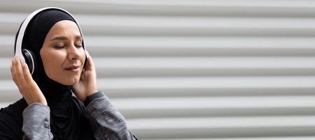 Satisfied young islamic lady in hijab in wireless headphones enjoy music on gray wall background