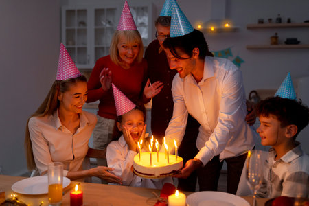 Adorable little girl getting birthday cake with lit candles from loving family, celebrating holiday at home in eveningの写真素材