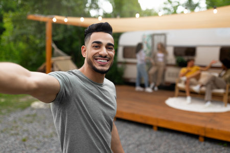 Happy Arab guy taking selfie near motorhome, resting on campsite with multiracial friends, enjoying...