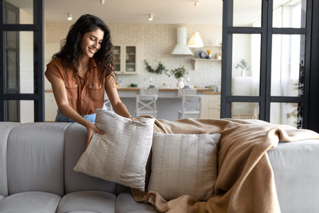 Lovely young woman putting soft pillows and plaid on comfy sofa, making her home cozy and warm, copy spaceの写真素材