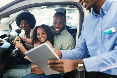 Black family testing new car, salesman showing them purchase or lease agreement on digital tablet at...