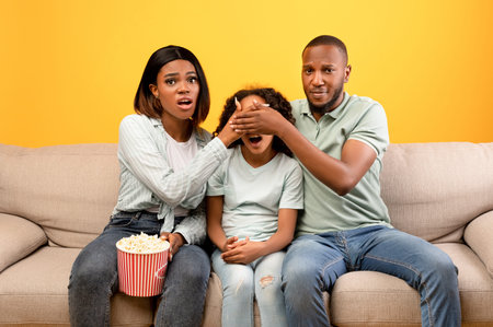 Scary moment in horror movie. African american parents closing daughters eyes while watching TV, sitting on sofaの写真素材
