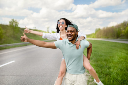 African American guy giving his girlfriend piggyback ride on highway, hitchhiking for car outdoorsの写真素材